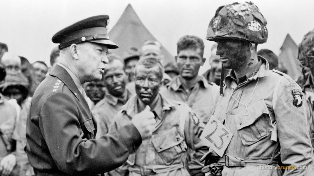 General Dwight D. Eisenhower speaks with a soldier of the 101st Airborne Division, whose face is painted for a night combat mission, while other paratroopers stand in the background.