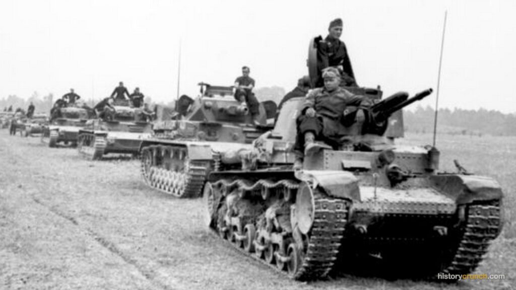 A column of German Panzer tanks advances along a dirt road during World War II, with crew members riding on top of the vehicles and infantry soldiers visible on the left side of the road.